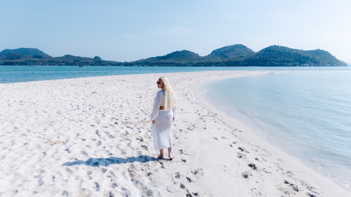 A woman with long blonde hair wearing a white dress stands on a sandy beach, facing the camera with sunglasses, with a serene sea and hills in the background.