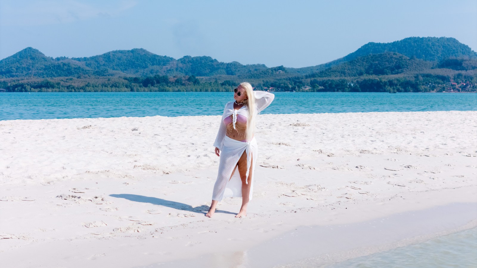 A woman with long blonde hair stands on a sandy beach, wearing a sheer white cover-up and sunglasses, with a backdrop of blue water and hills.