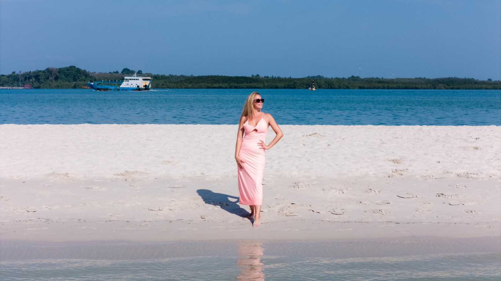 A woman in a pink dress stands on a sandy beach, with clear blue water and a boat in the background.