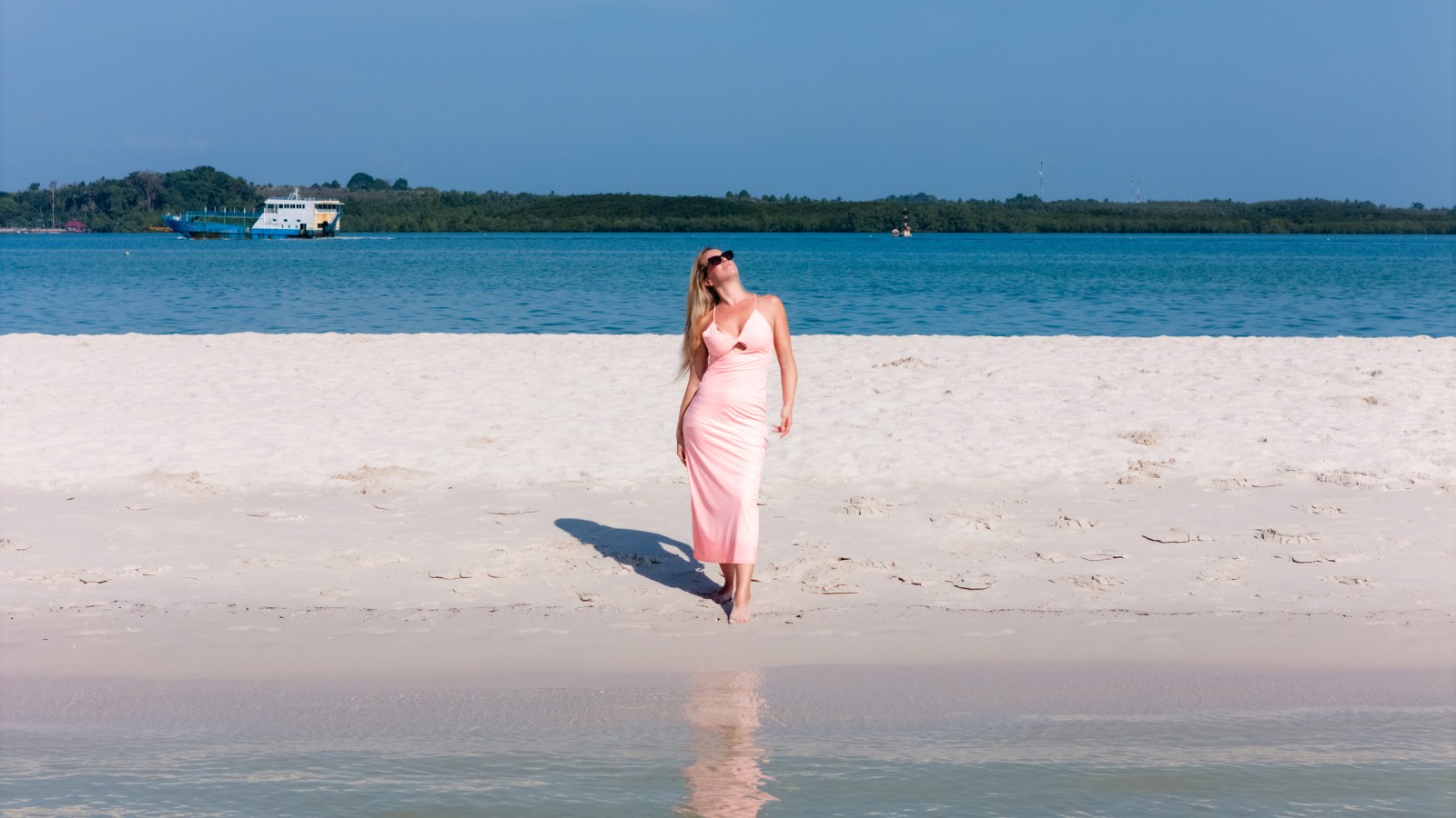 A woman in a pink dress walking on a sandy beach near calm turquoise waters with a boat in the background.