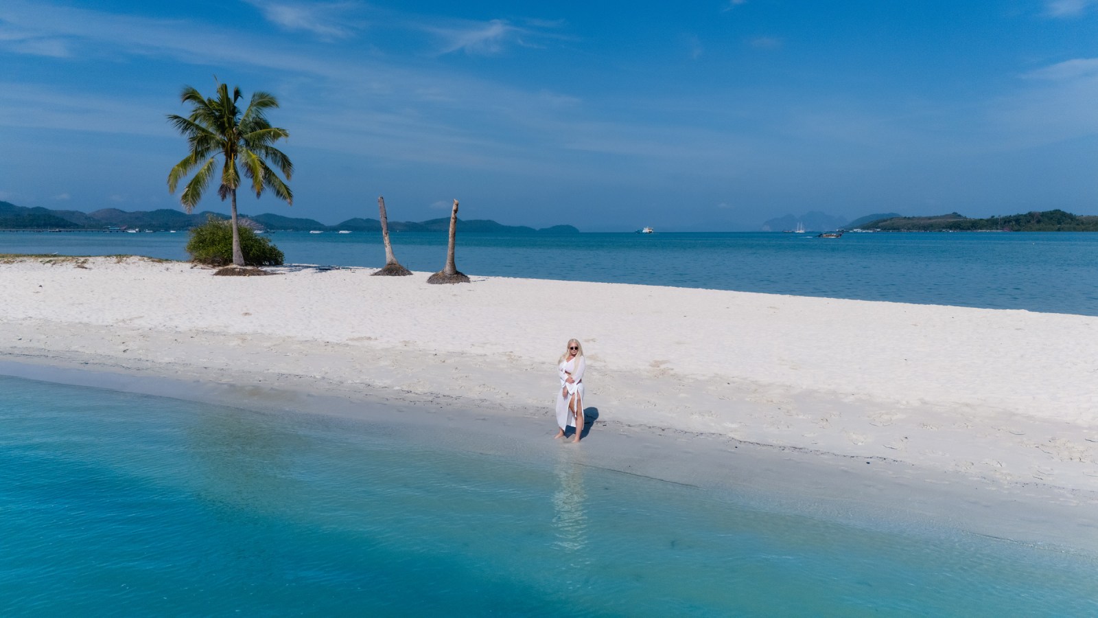 A woman standing barefoot on a sandy beach by the turquoise water, with palm trees and distant islands under a clear blue sky.