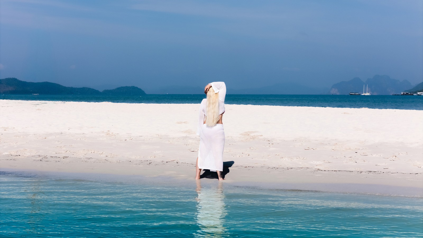 A woman in a white dress stands by the water on a sandy beach, with a backdrop of blue sea and distant islands under a clear sky.