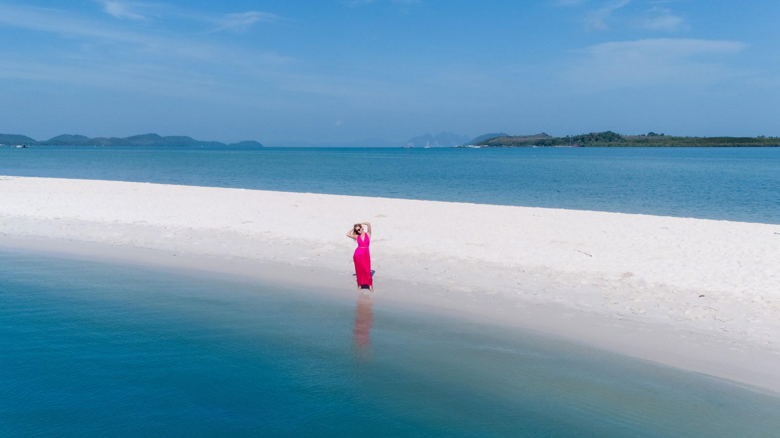 A woman in a pink dress stands on a pristine white sandy beach, with turquoise water and distant mountains in the background under a clear blue sky.