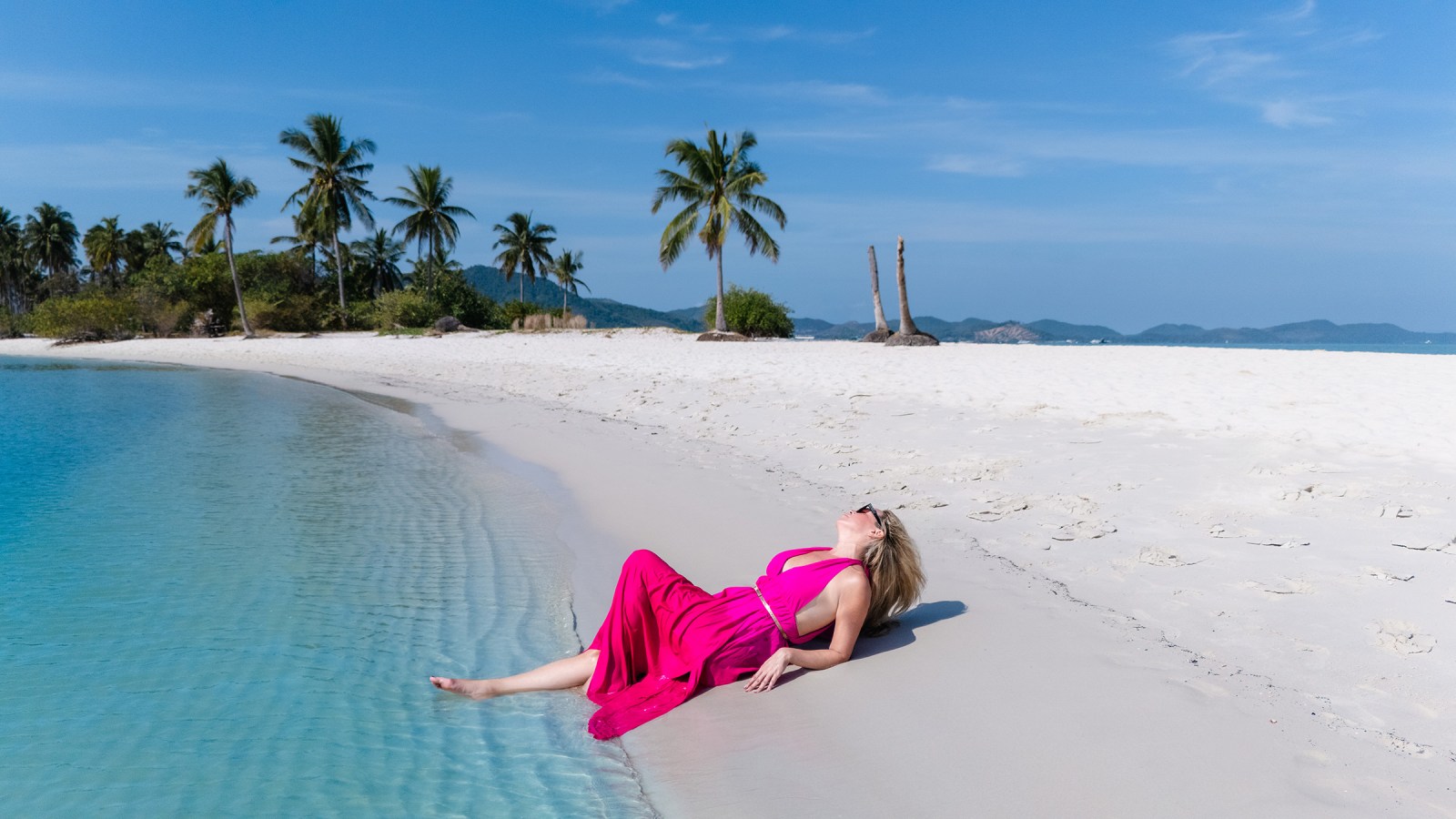 A woman in a pink dress lounging on a sandy beach near clear turquoise water, with palm trees and distant hills in the background.
