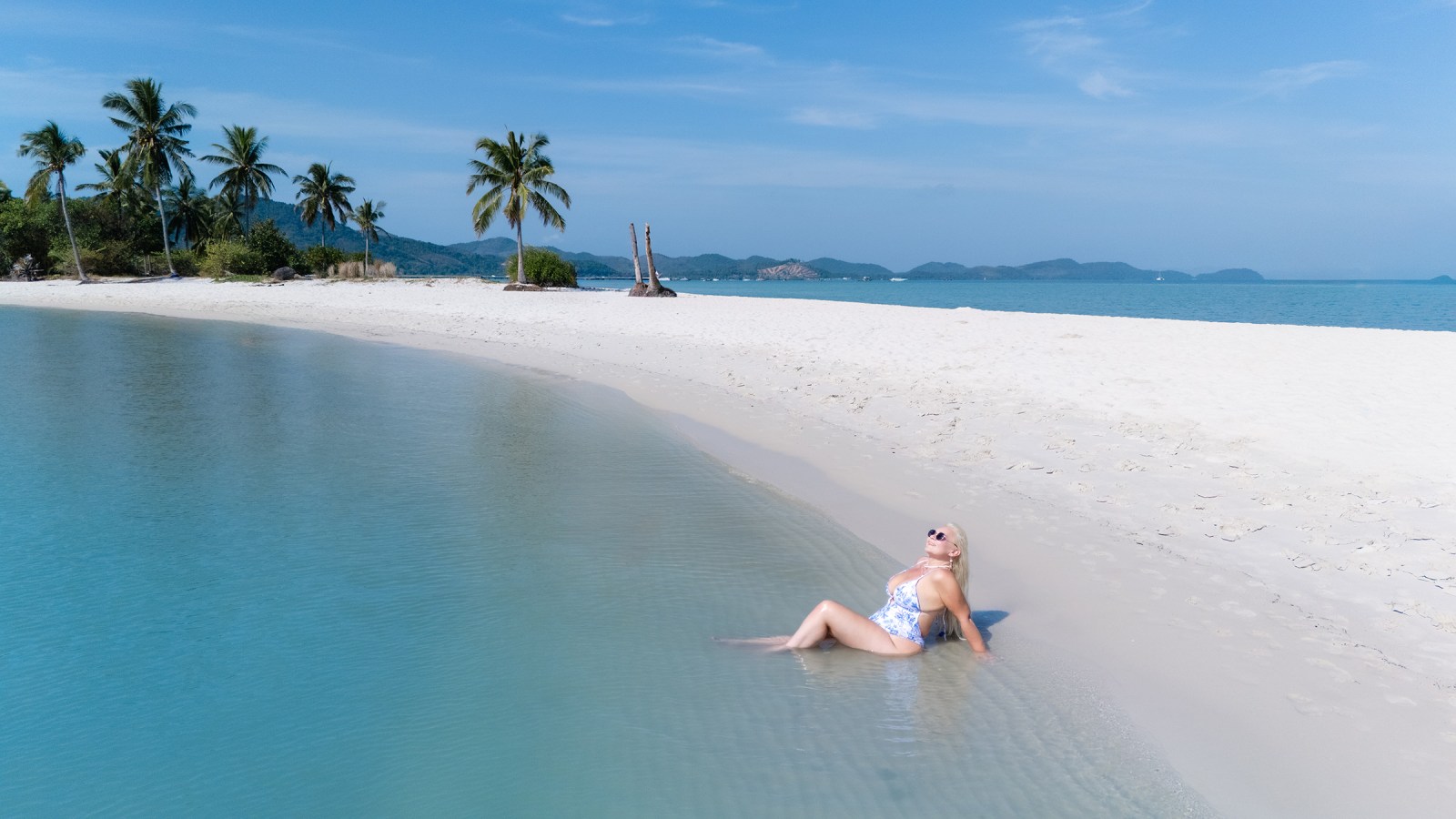 A woman relaxing on the beach in shallow water, surrounded by palm trees and a scenic coastline under a clear blue sky.
