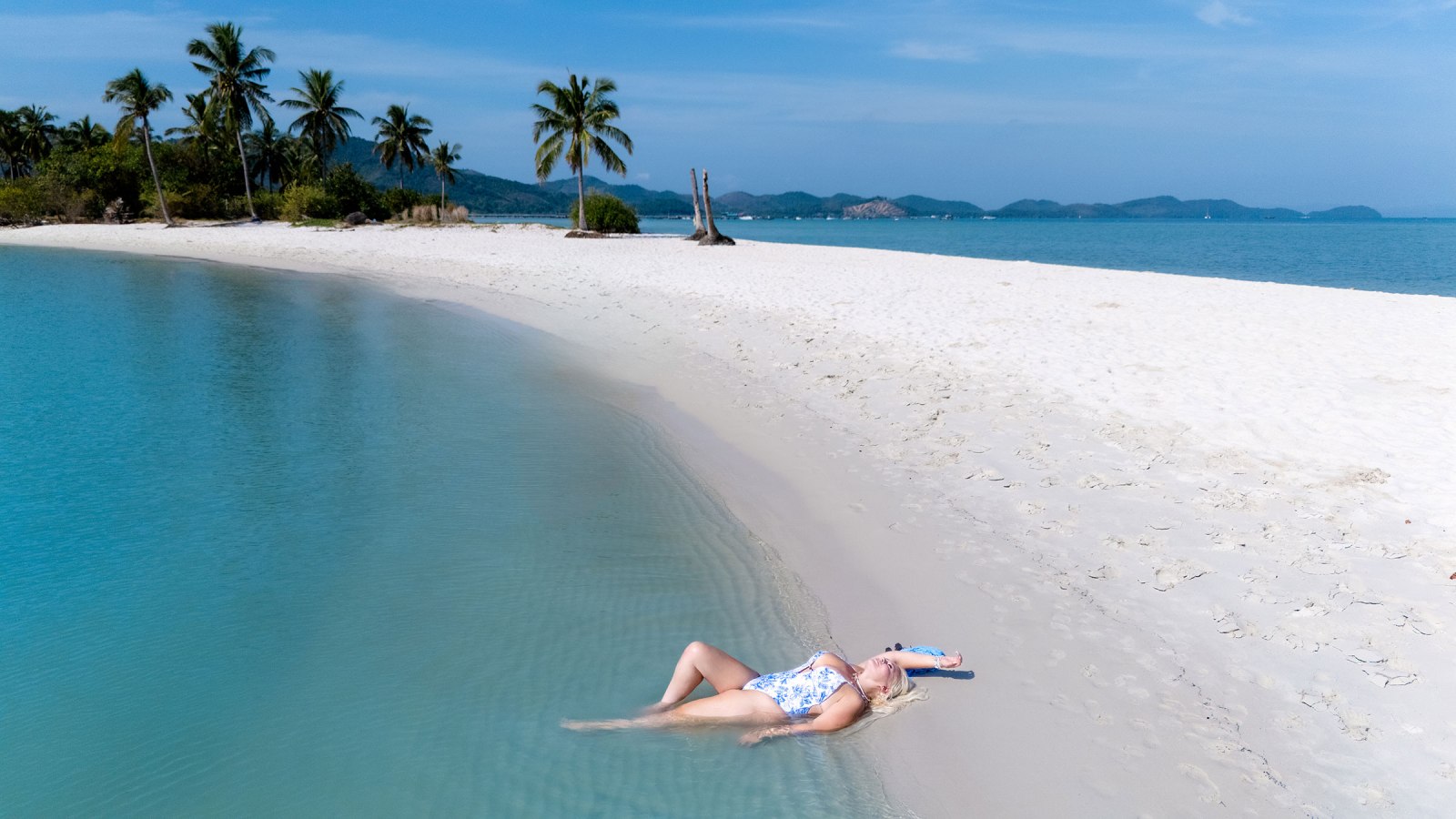 A person relaxing on a sandy beach by a turquoise lagoon, surrounded by palm trees and islands in the background.