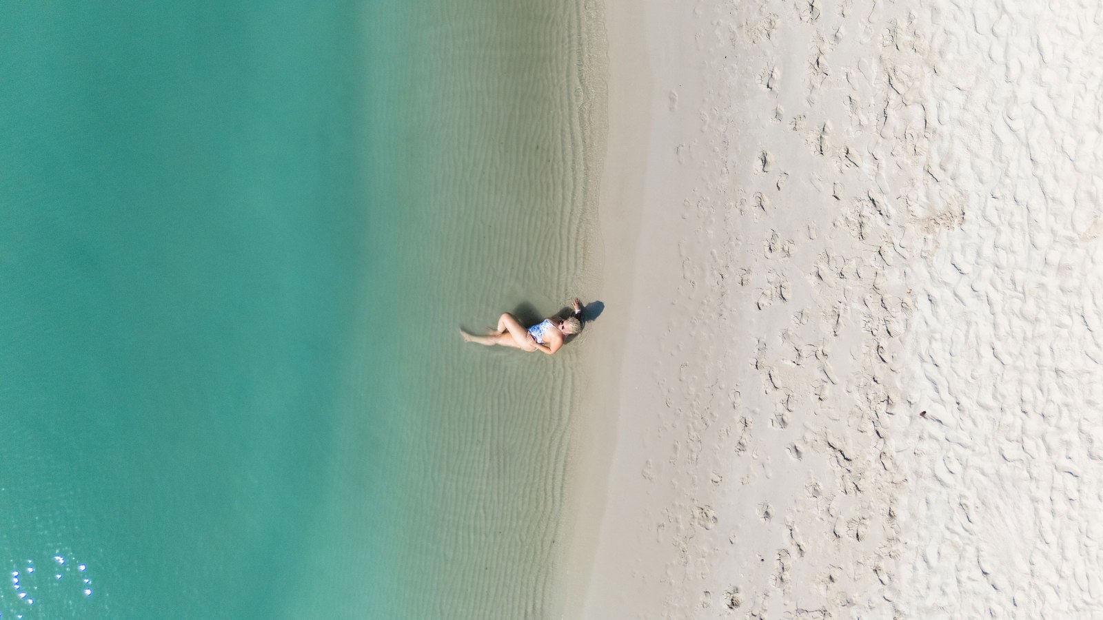 Aerial view of a person lying on a sandy beach by clear turquoise water.
