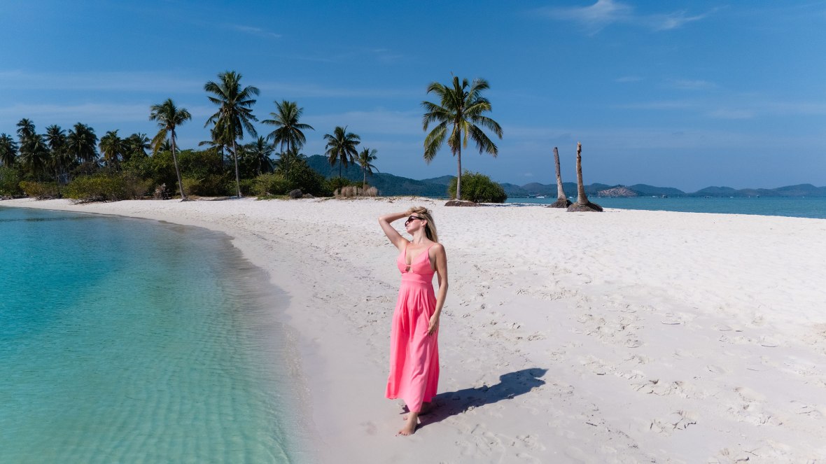 A woman in a pink dress standing on a sandy beach with palm trees and clear blue water in the background.