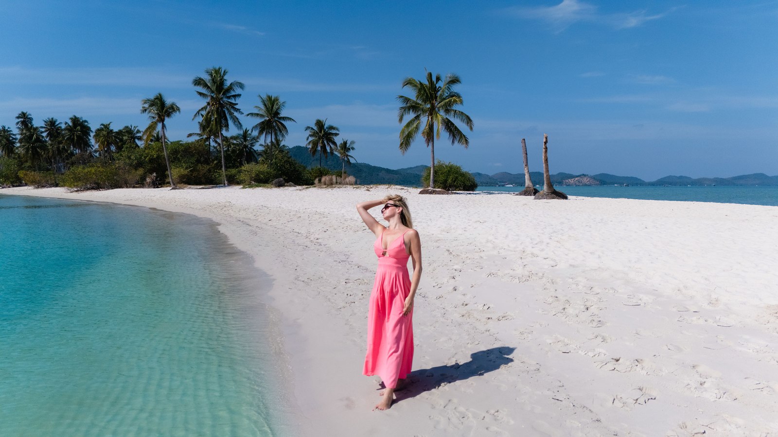 A woman in a pink dress stands on a sandy beach near clear turquoise water, with palm trees and mountains in the background under a blue sky.