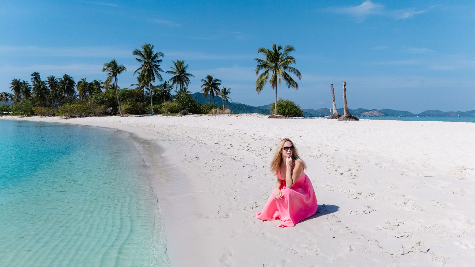 A woman in a pink dress sitting on a white sandy beach beside clear blue water, with palm trees and mountains in the background.