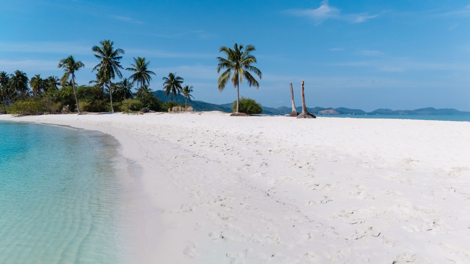 A serene beach scene with white sand, clear turquoise water, and tall palm trees under a blue sky.