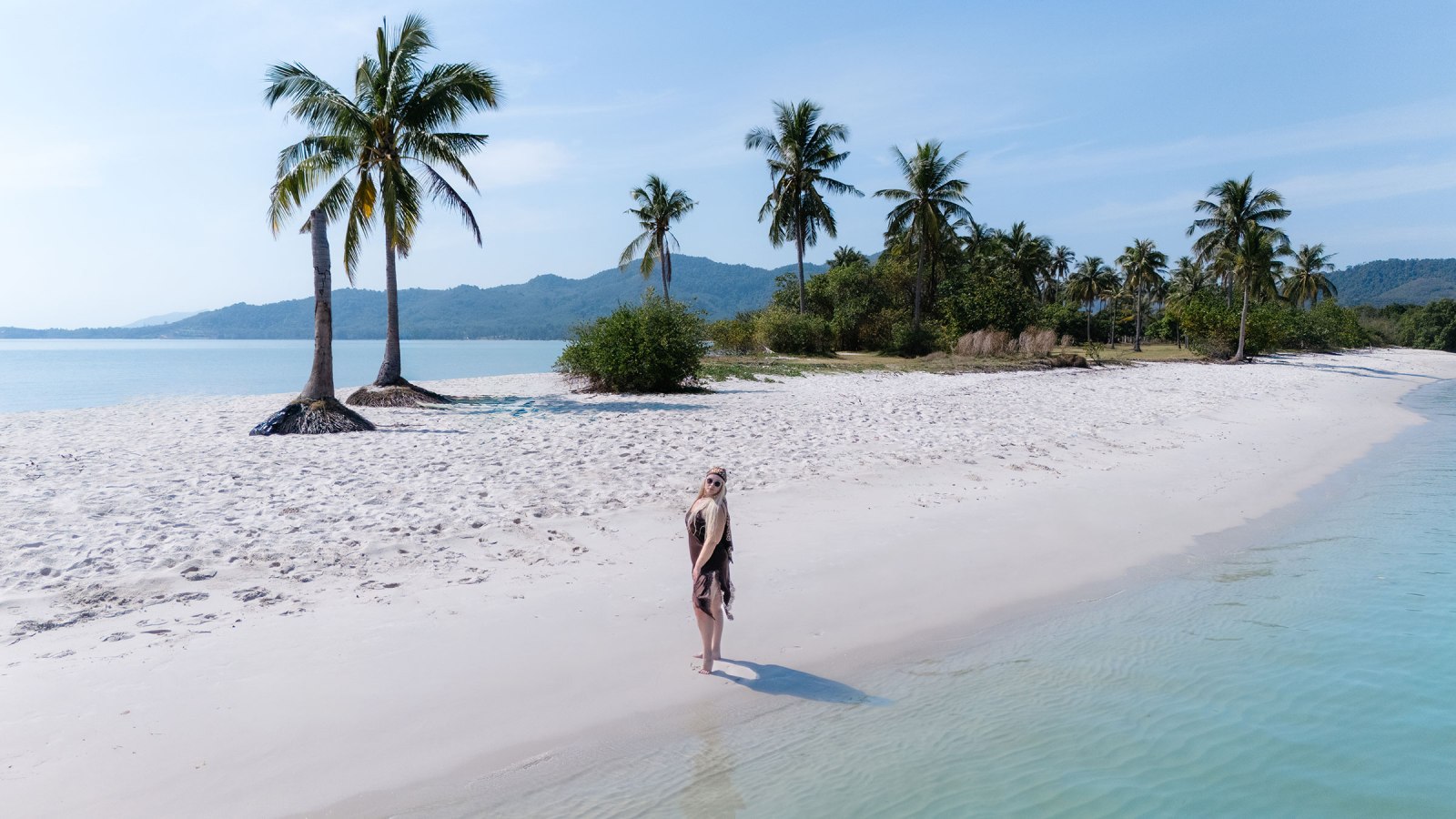 A woman standing on a sandy beach next to calm turquoise waters, surrounded by palm trees and distant hills under a clear sky.