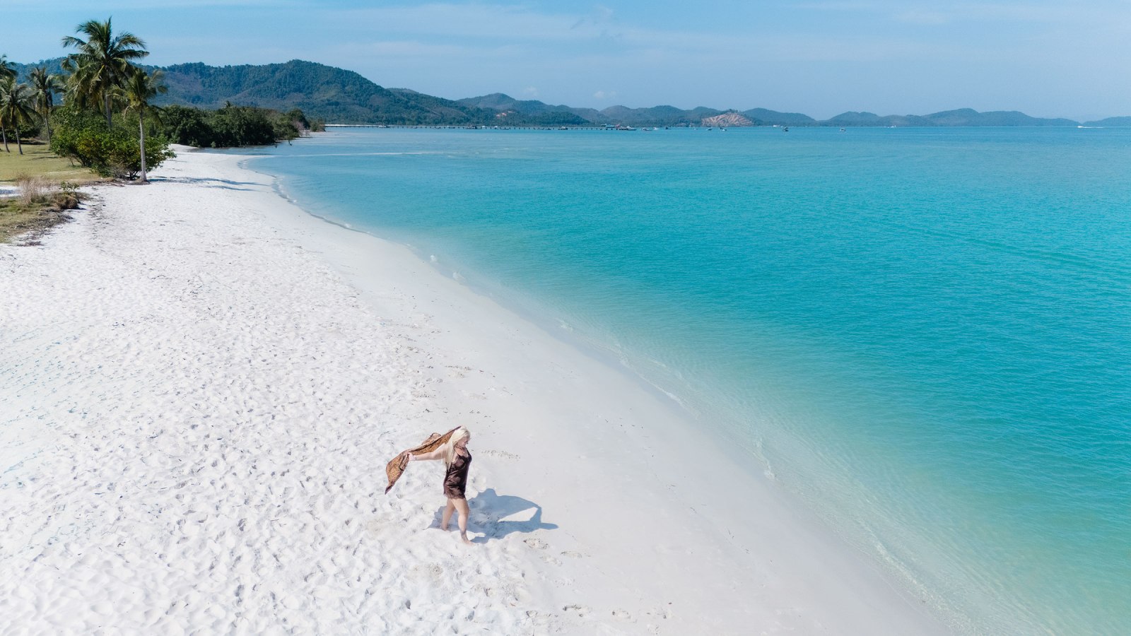 A person standing on a sandy beach with turquoise water and lush hills in the background, joyfully waving a scarf.