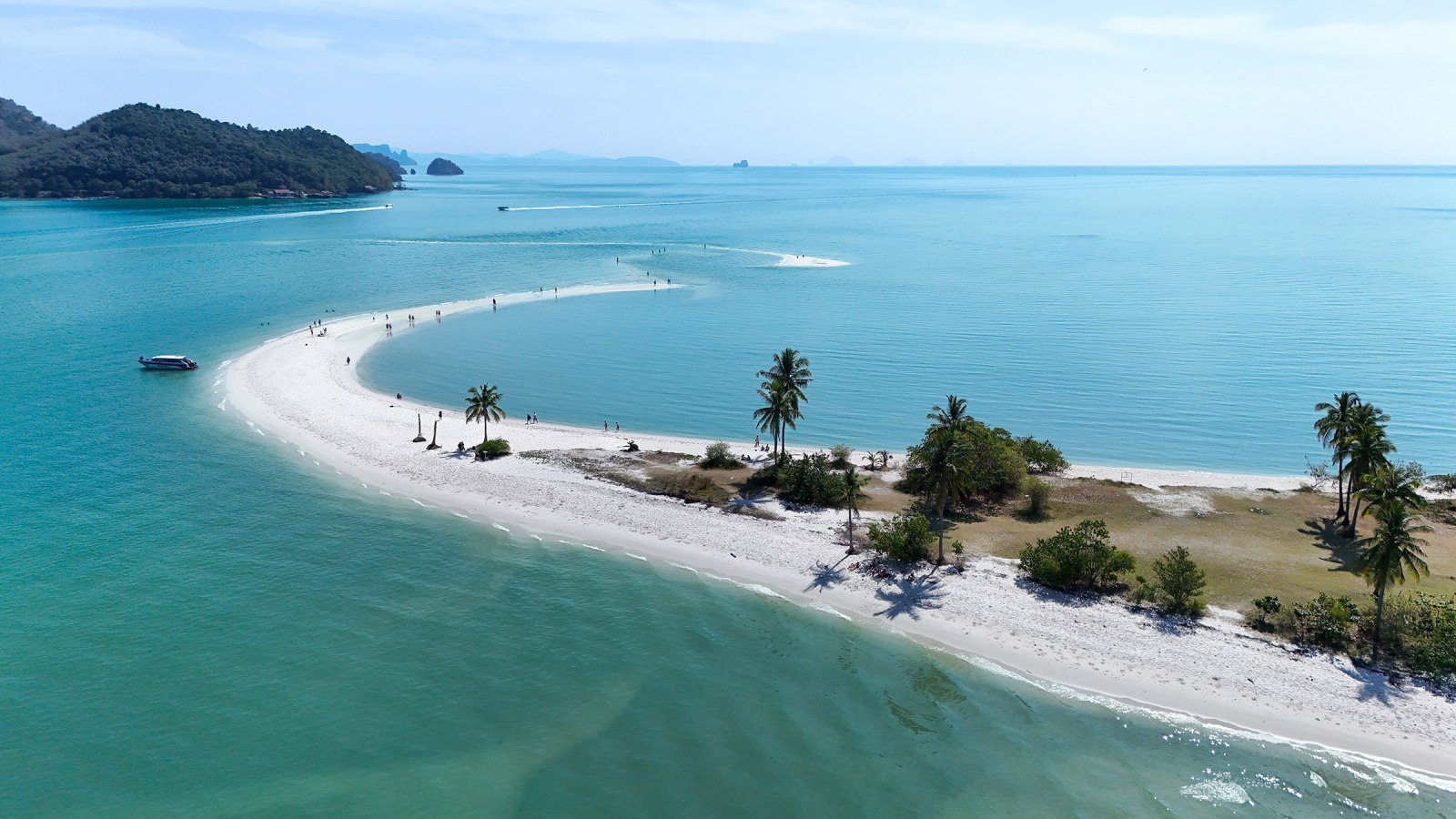 Aerial view of a picturesque beach with white sand and clear turquoise water, featuring palm trees and people walking along a curved shoreline.