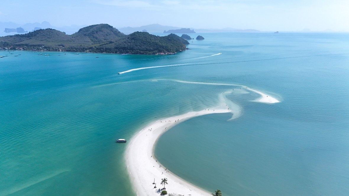 Aerial view of a white sandy beach forming a curved peninsula surrounded by turquoise waters and lush green islands in the background.
