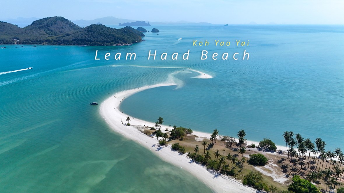 Aerial view of Leam Haad Beach on Koh Yao Yai, featuring a curved sandbar, turquoise waters, and lush green hills in the background.