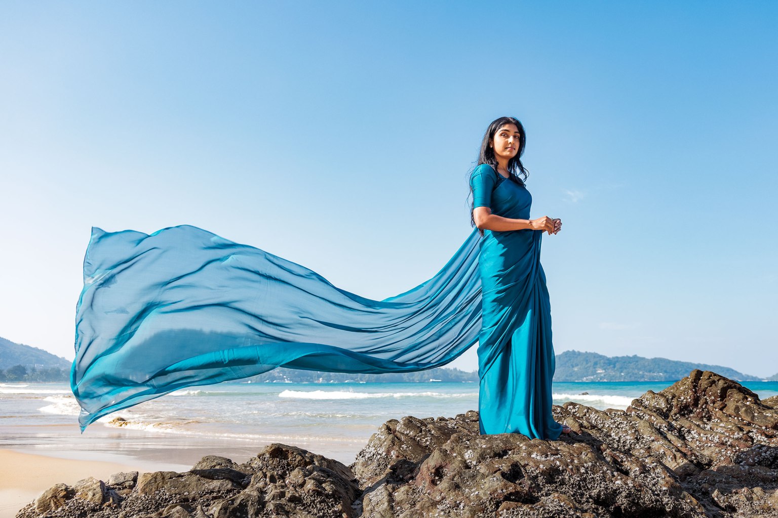 A woman standing on rocks by the beach, wearing a flowing turquoise sari that billows in the wind, with a clear blue sky and ocean in the background.