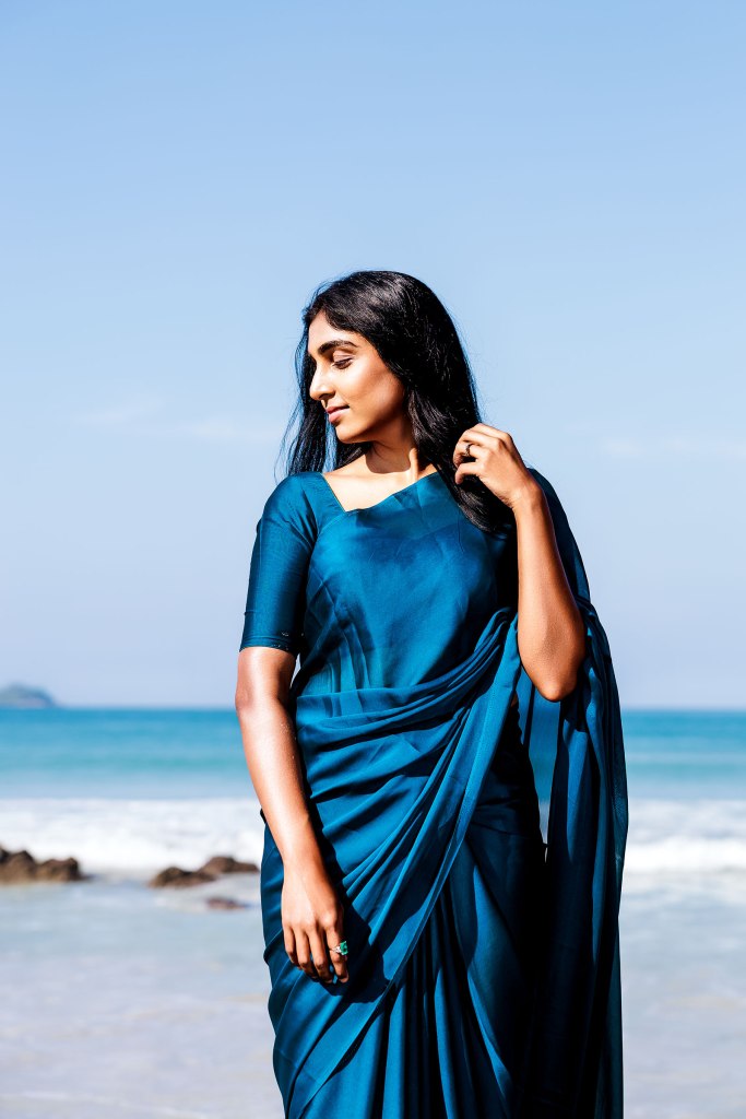 A woman wearing a blue saree stands on a beach with the ocean in the background, her hair flowing and hand gently touching her hair.