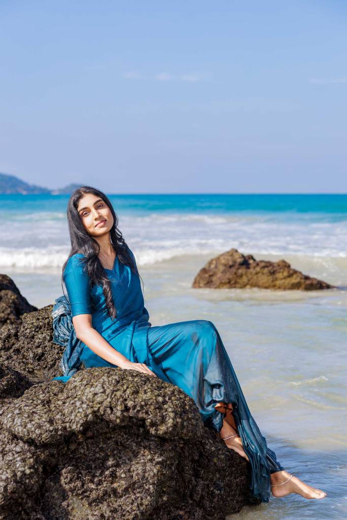 A smiling woman in a teal dress sits on a rocky beach with the ocean in the background on a sunny day.