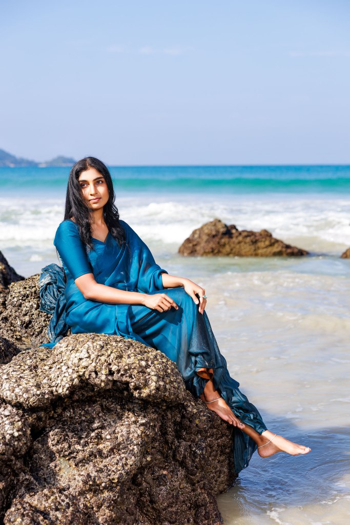 A woman in a blue saree sitting on a rocky beach, with waves crashing in the background and a clear blue sky overhead.