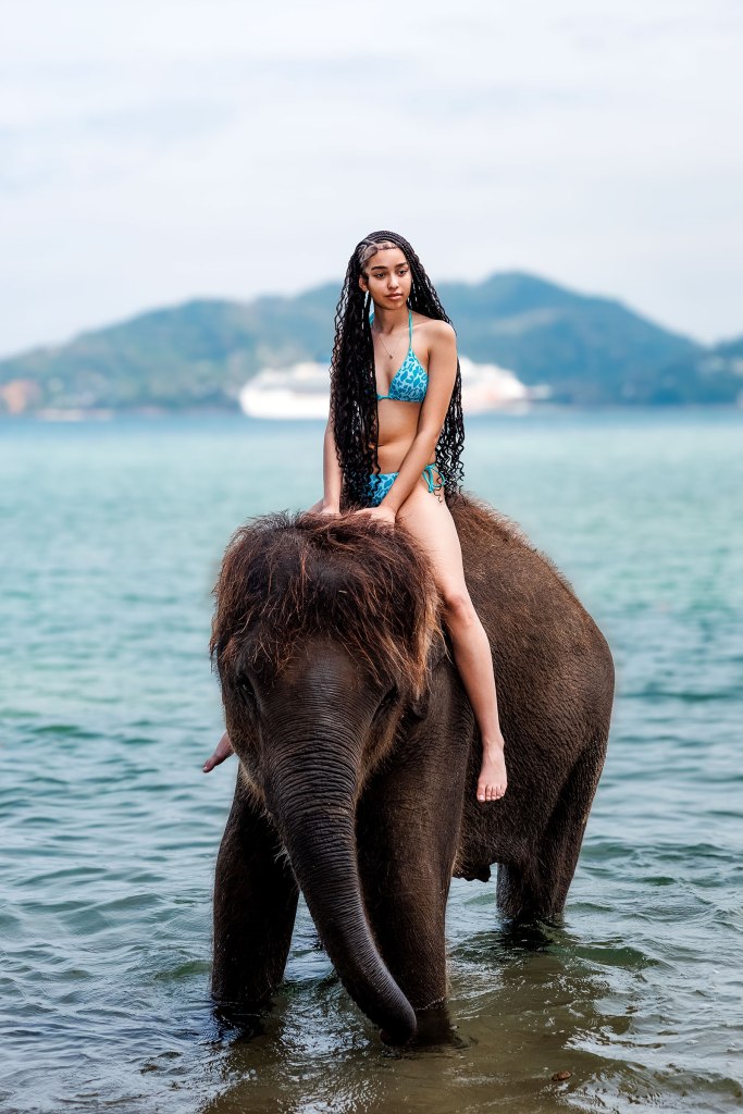 A women in a blue bikini sitting atop an elephant in shallow water, with mountains in the background for photo shooting in phuket