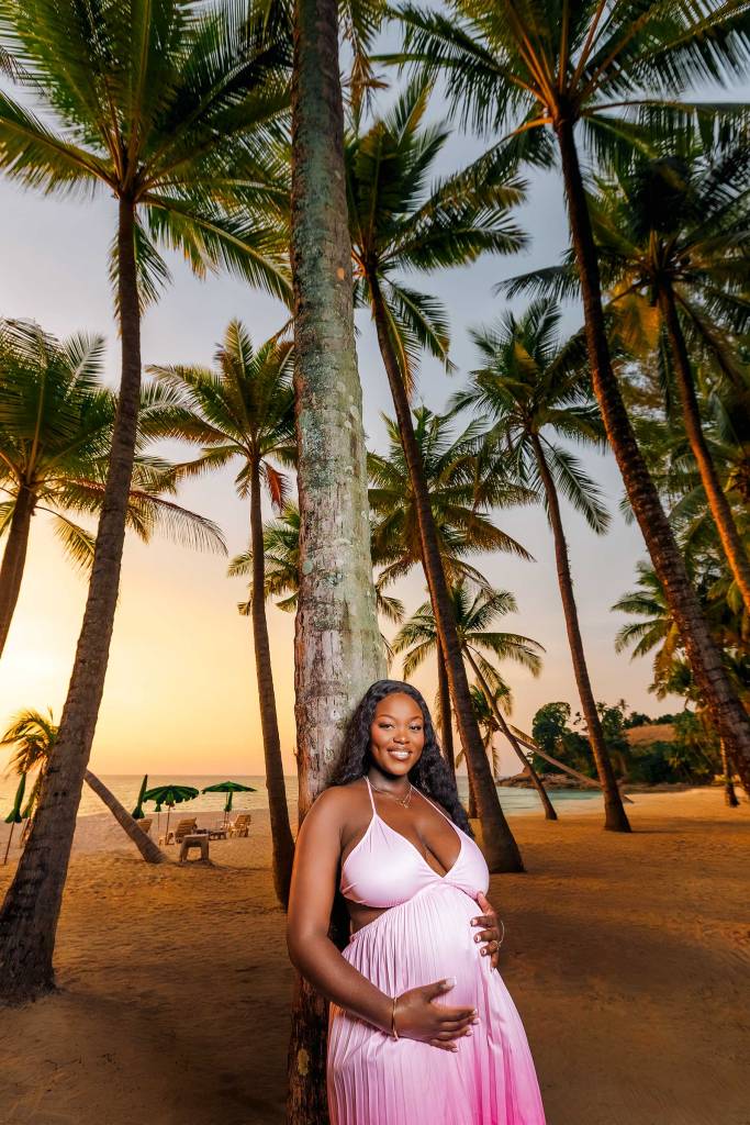 A pregnant woman in a pink dress stands beside a palm tree on a sandy beach during sunset, with palm trees and beach chairs in the background.