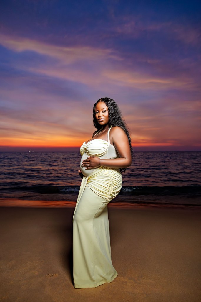 A pregnant woman in a yellow dress stands on the beach at sunset, with the ocean in the background and colorful clouds in the sky.
