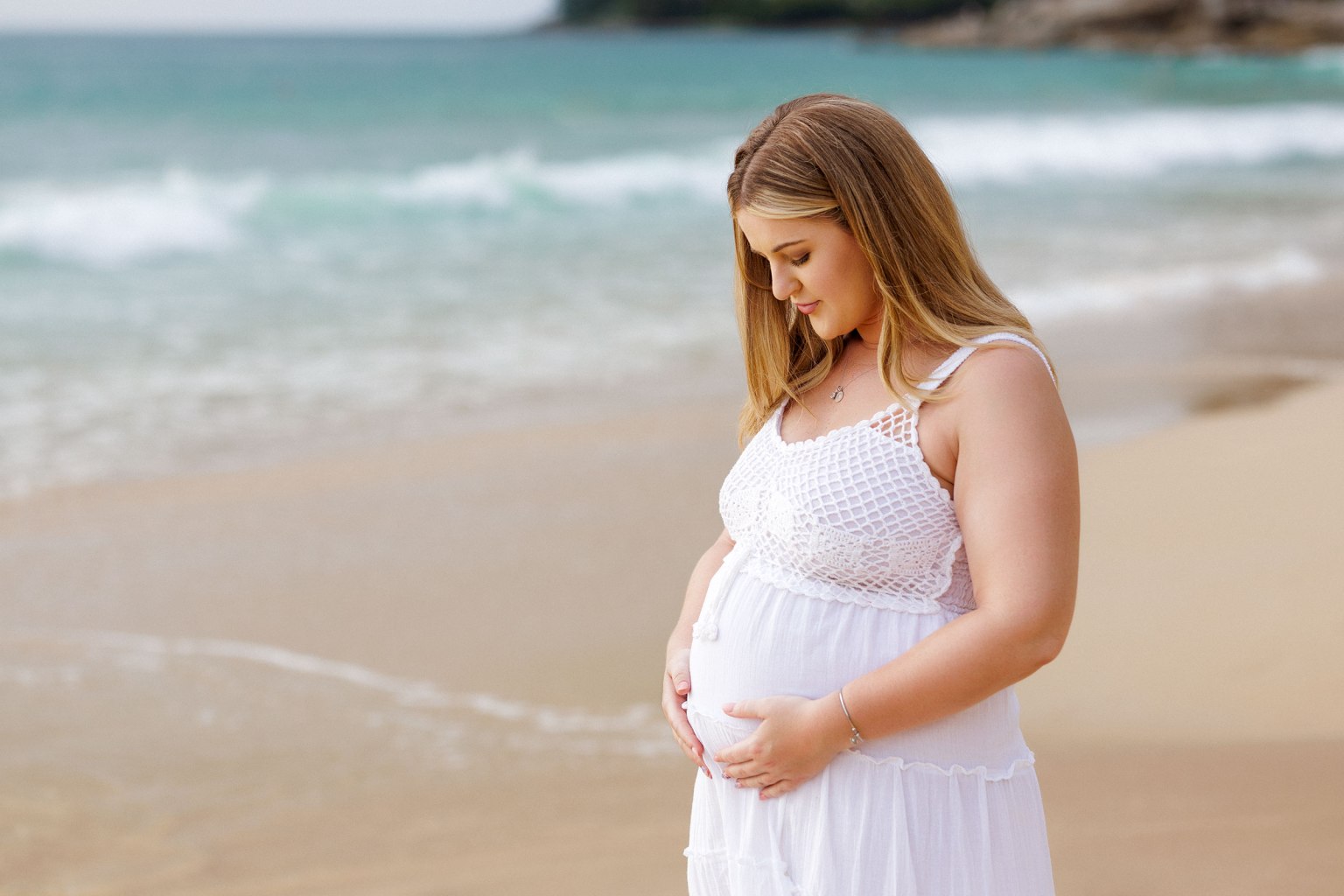 Pregnant woman in a white dress gently cradling her belly while standing on a beach with gentle waves in the background.