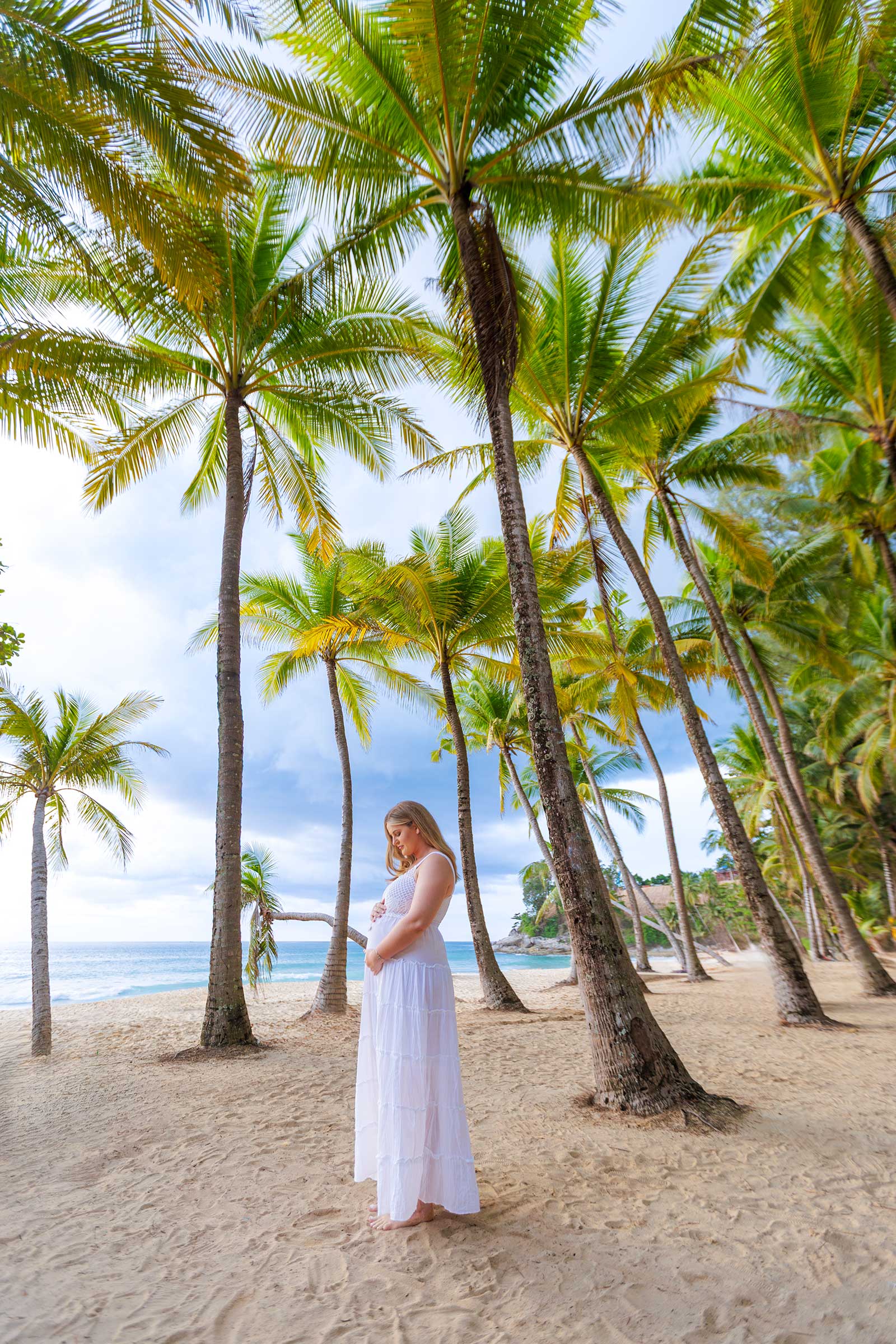 A pregnant woman in a white dress standing on a sandy beach surrounded by tall palm trees, with the ocean in the background under a cloudy sky.