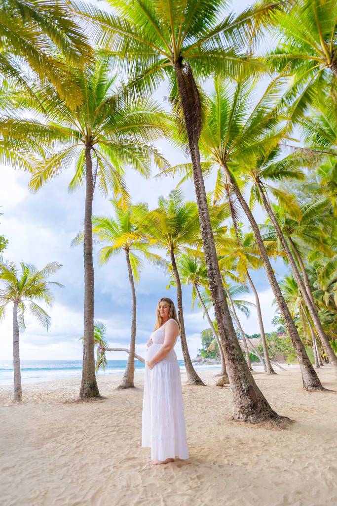 A pregnant woman in a white dress standing on a sandy beach, surrounded by tall palm trees and a serene ocean view under a cloudy sky.