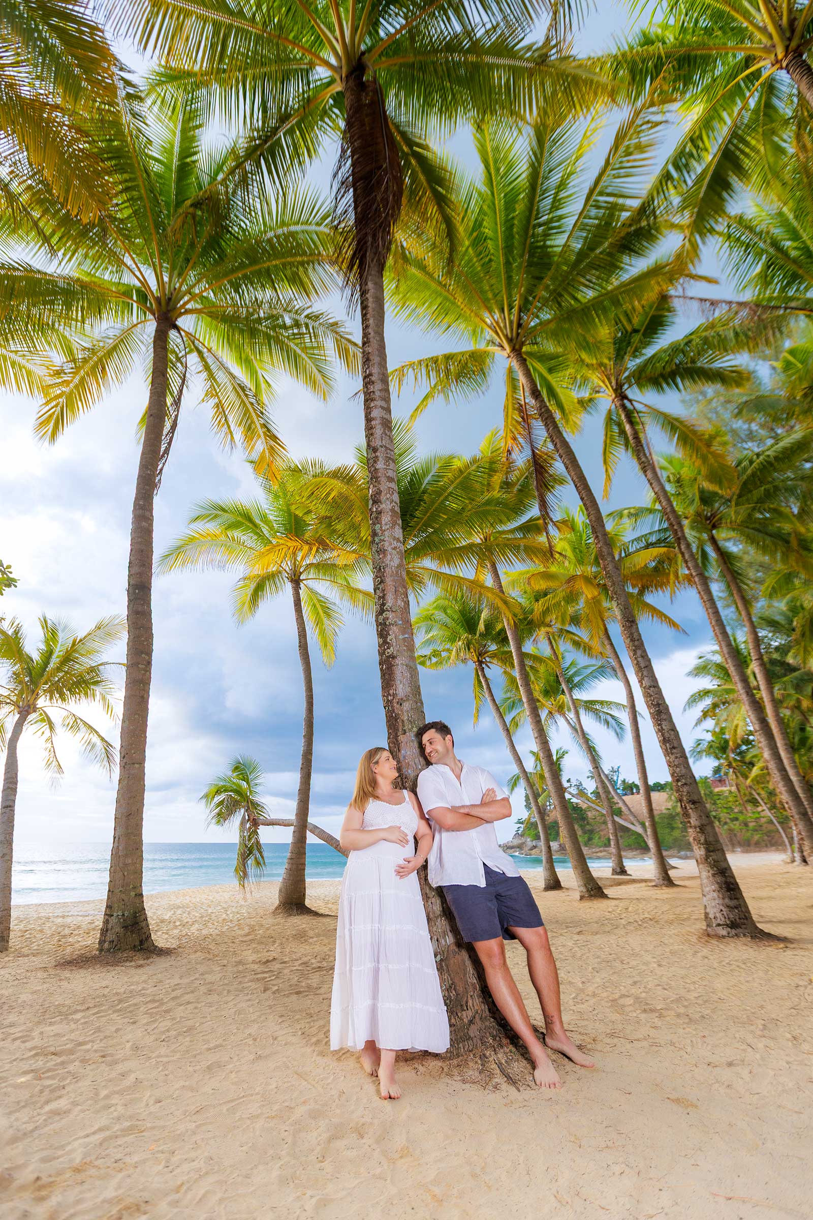 A couple standing under palm trees on a sandy beach, with the ocean in the background. The woman is wearing a white dress, and the man is dressed in a white shirt and shorts. They are smiling and looking at each other.