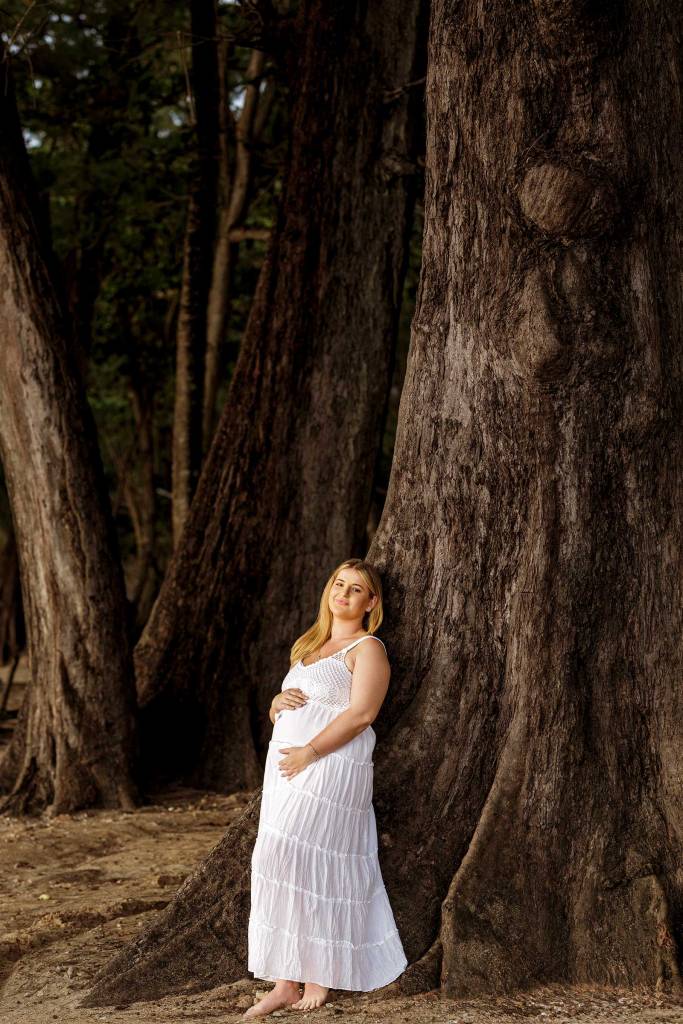 A pregnant woman in a white dress stands barefoot next to a large tree, embracing her baby bump in a serene outdoor setting.
