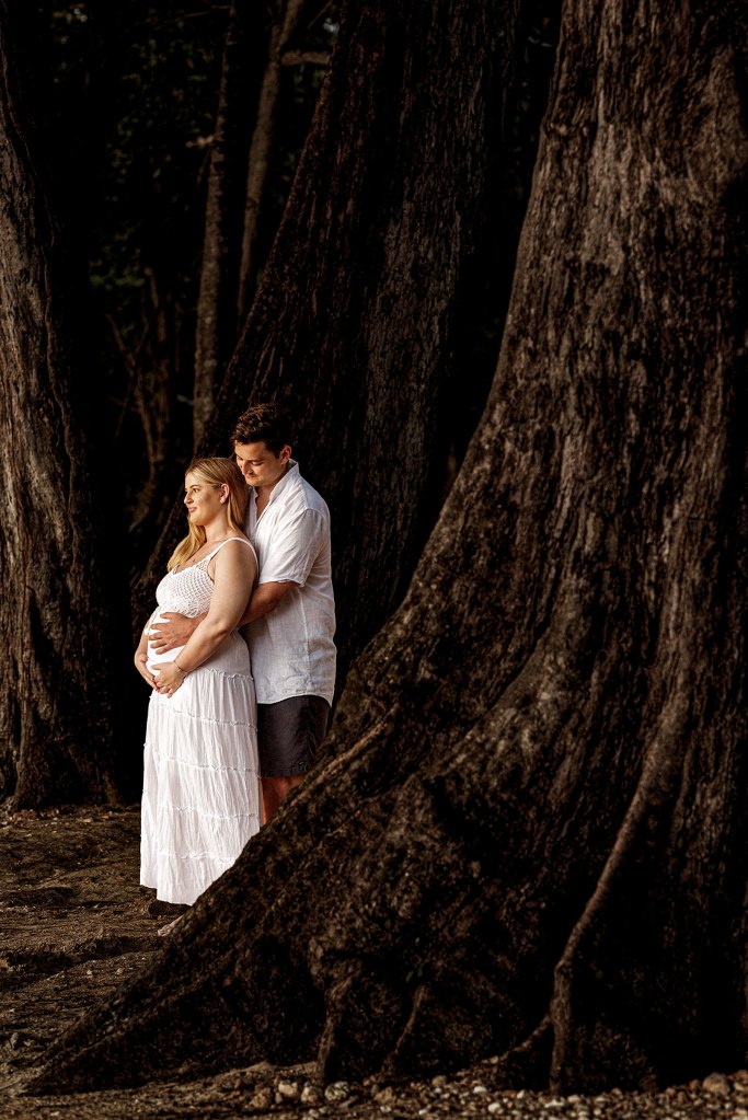 A pregnant woman in a white dress stands near a tree, lovingly embraced by her partner, with dark trees in the background.