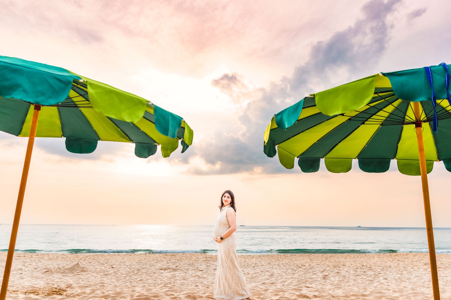 A woman in a flowing dress stands on a sandy beach between two colorful umbrellas, with the ocean and sunset in the background.