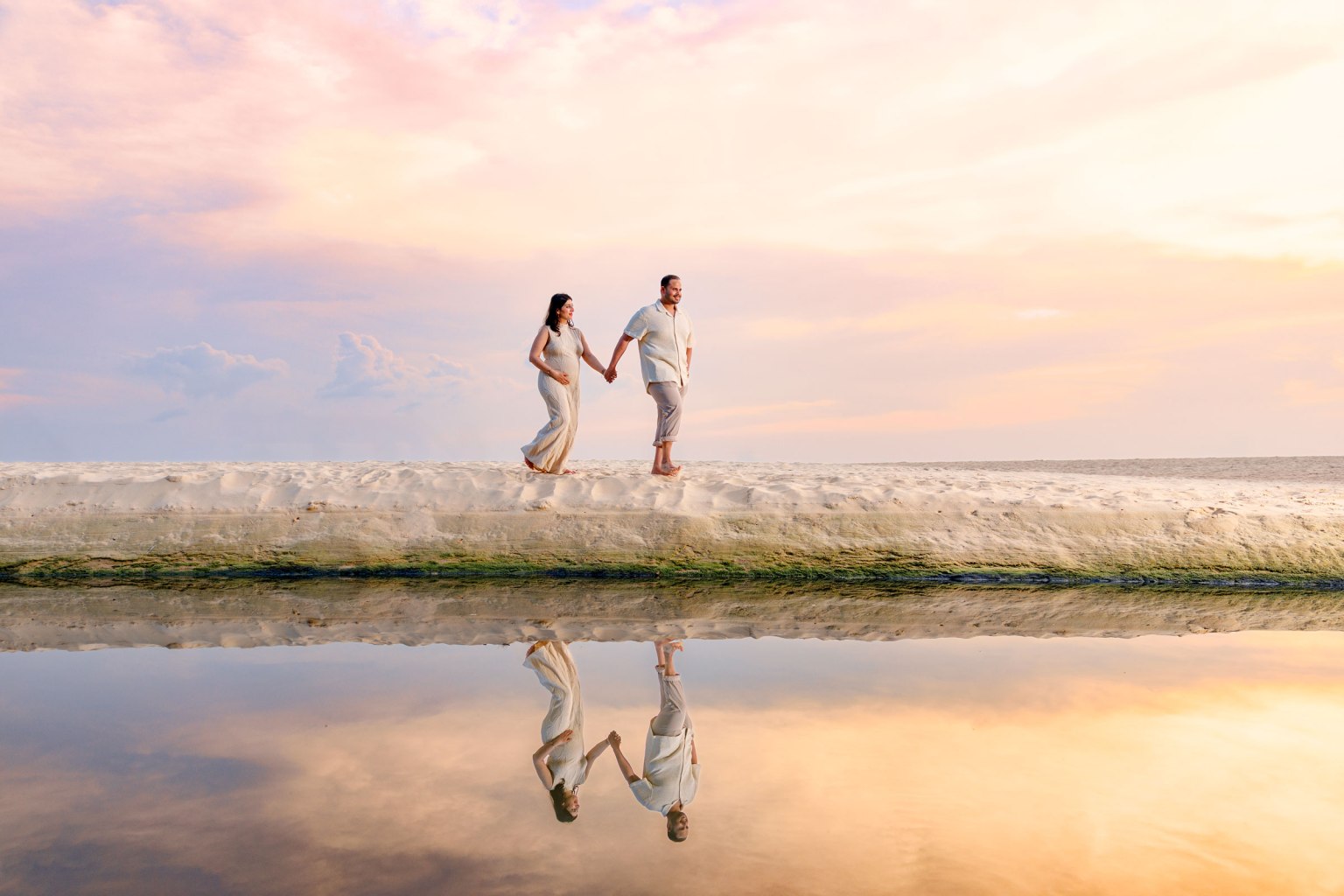 A couple at karon beach holding hands while walking on a sandy beach at sunset, with their reflections visible in the calm water beside them.