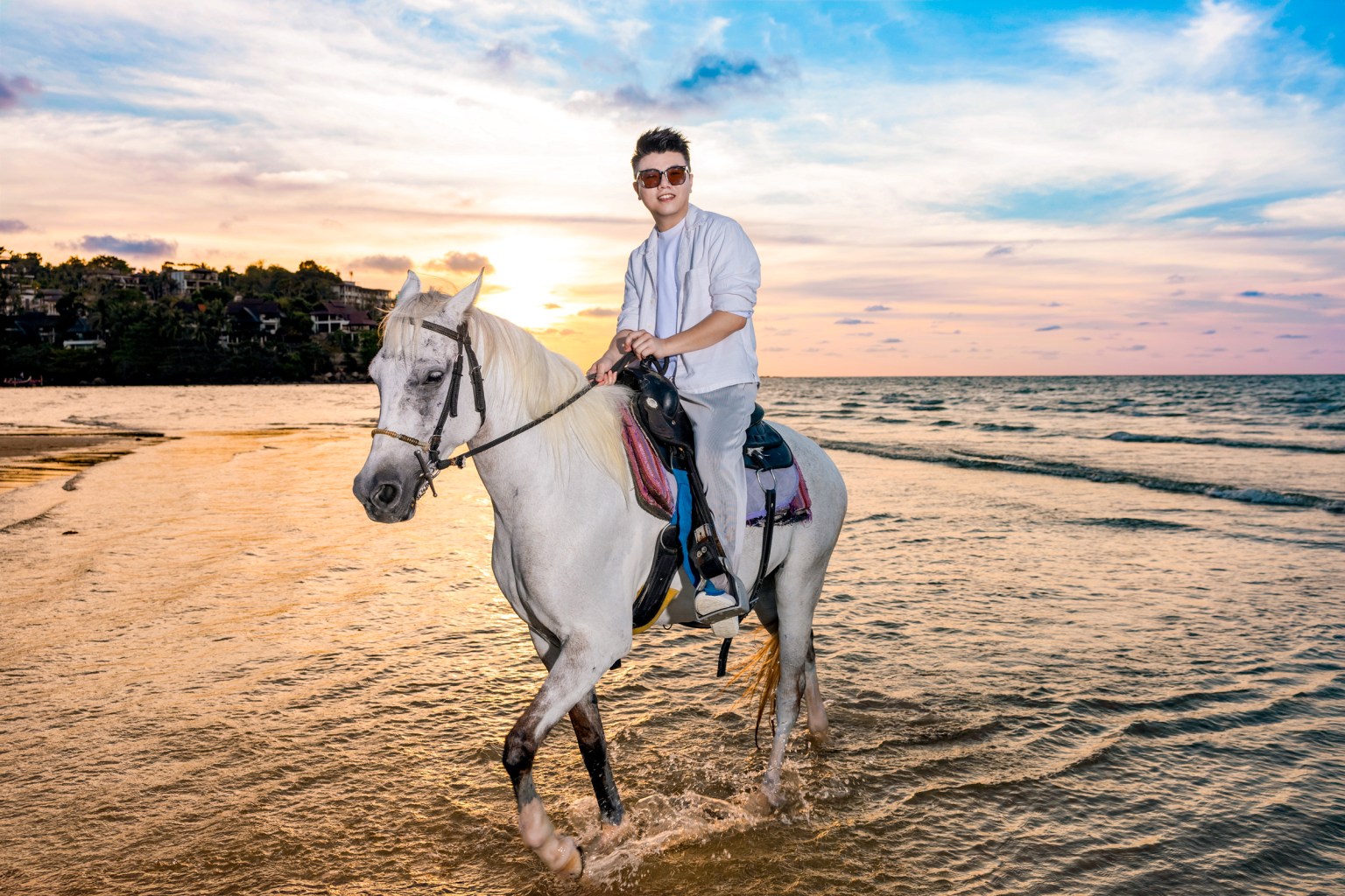 A young person riding a white horse along the shoreline at sunset, with ocean waves in the background and a colorful sky.