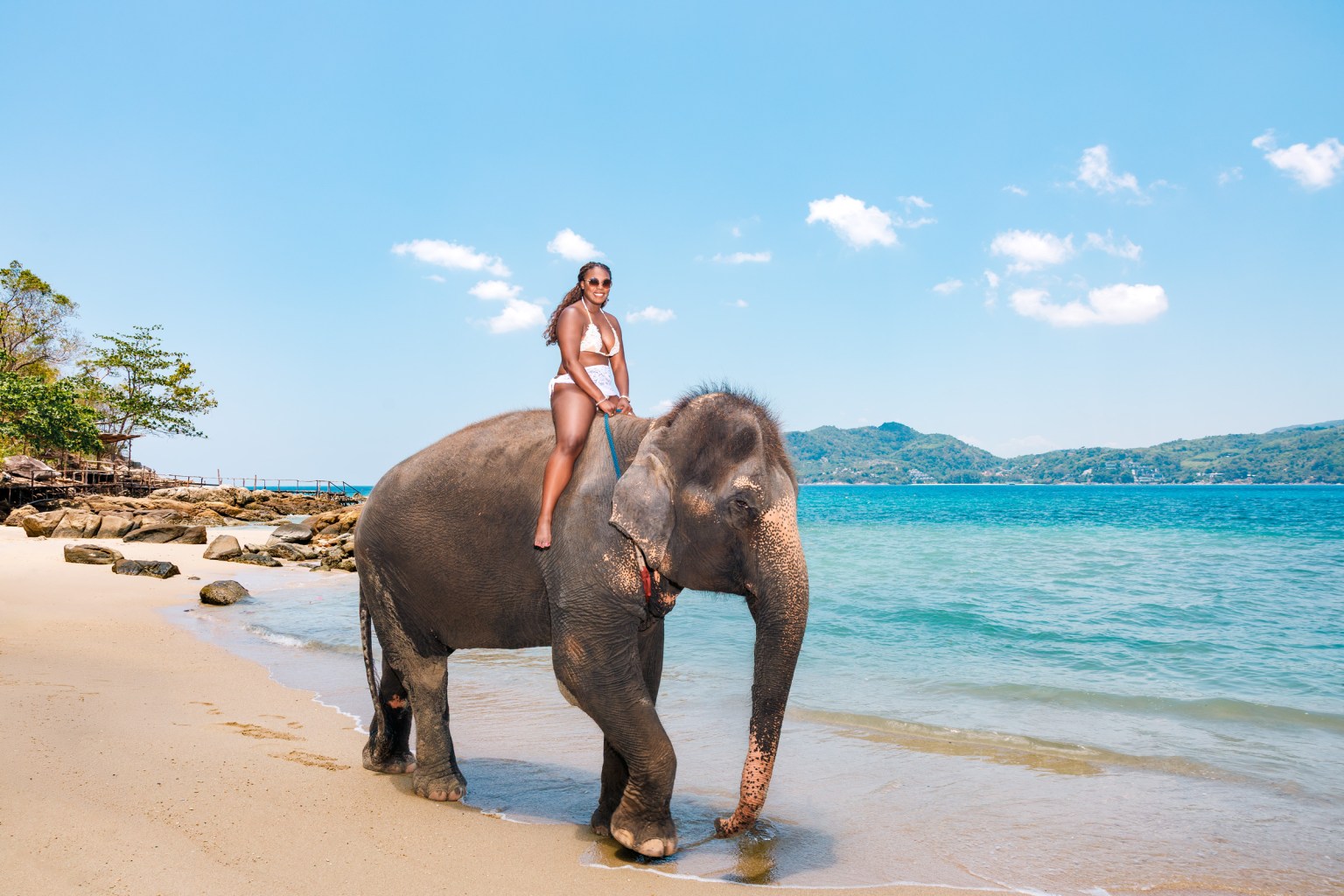 A woman in a bikini riding an elephant along a sandy beach with clear blue water and a scenic background.