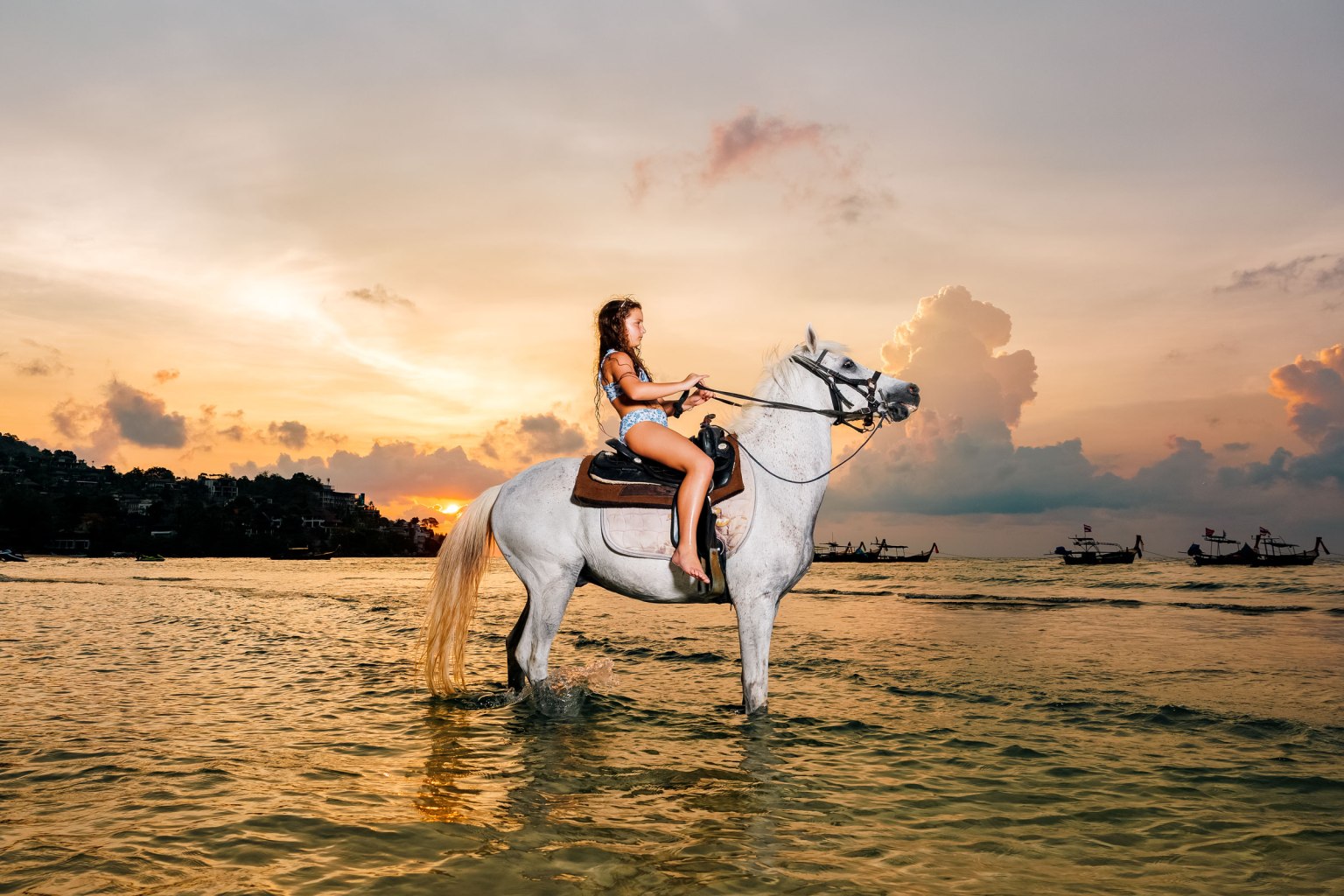 A young girl in a swimsuit riding a white horse in shallow water during sunset at kamala beach phuket, with long tail boats in the background.